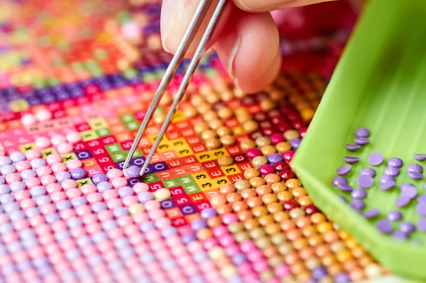 girl collect diamond painting. diamond embroidery with a tweezers. acrylic rhinestones. close up, selective focus.
